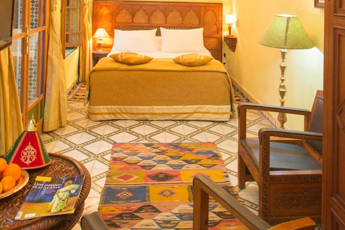 A hotel room with a bed, a chair, a table with oranges and a book, a patterned rug, and a chandelier.
