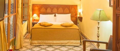 A hotel room with a bed, a chair, a table with oranges and a book, a patterned rug, and a chandelier.