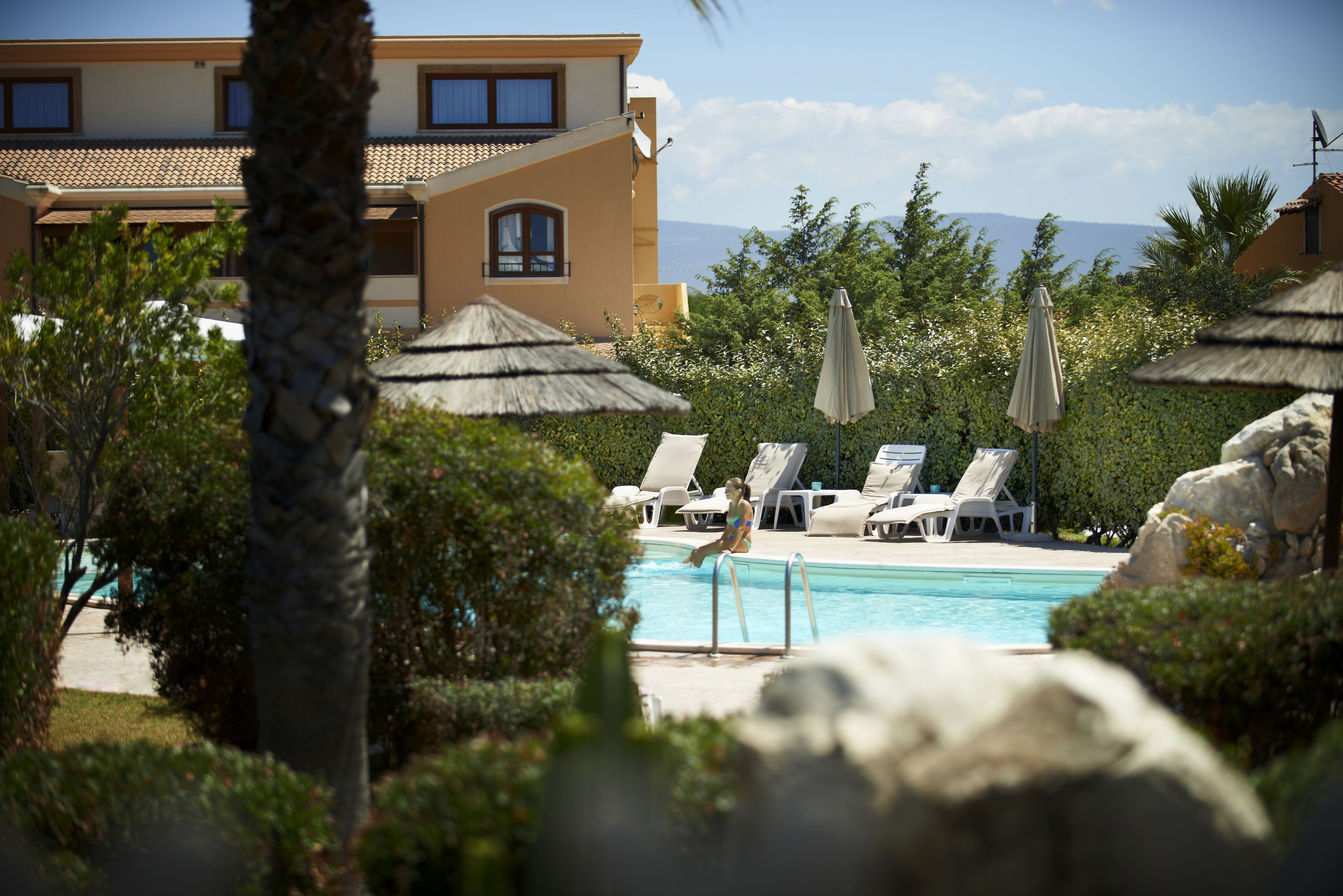 A pool area with lounge chairs and umbrellas, surrounded by greenery and a building in the background.