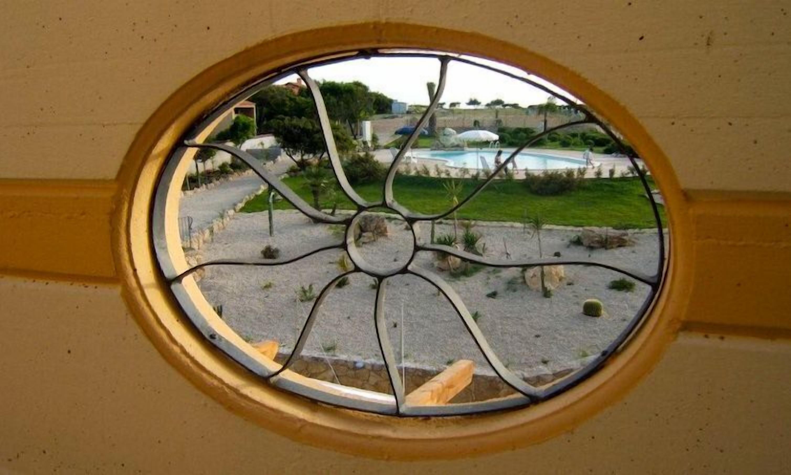 Vista a través d'una finestra circular d'un pati amb piscina, vegetació i un camí de pedra.