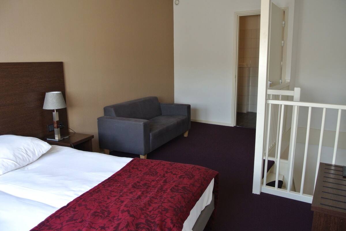 A hotel room with a bed, a chair, a nightstand with a lamp, and a small white crib.