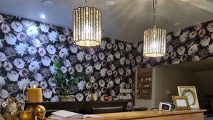 A reception desk with a floral wallpaper background, two pendant lights, a lit candle, and decorative wreaths. - Hotel Arcen (Arcen)