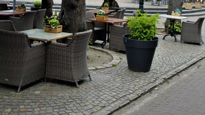 An outdoor cafe with wicker furniture, potted plants, and a brick building with a clock tower in the background. - Hotel Arcen (Arcen)