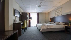 A hotel room with a bed, bedside table, lamp, TV, desk, chair, and a view of the outside. - Hotel Arcen (Arcen)