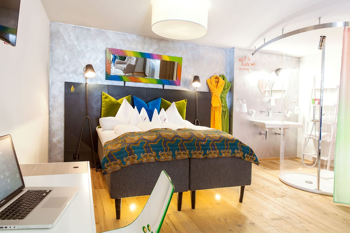 A modern bedroom with a bed, a laptop, a mirror, and a bathroom with a shower.