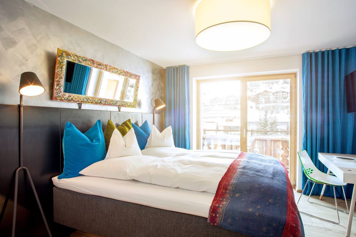 A neatly made bed with white linens and colorful pillows, a large mirror with a decorative frame, a desk with a chair, and a large window with curtains.