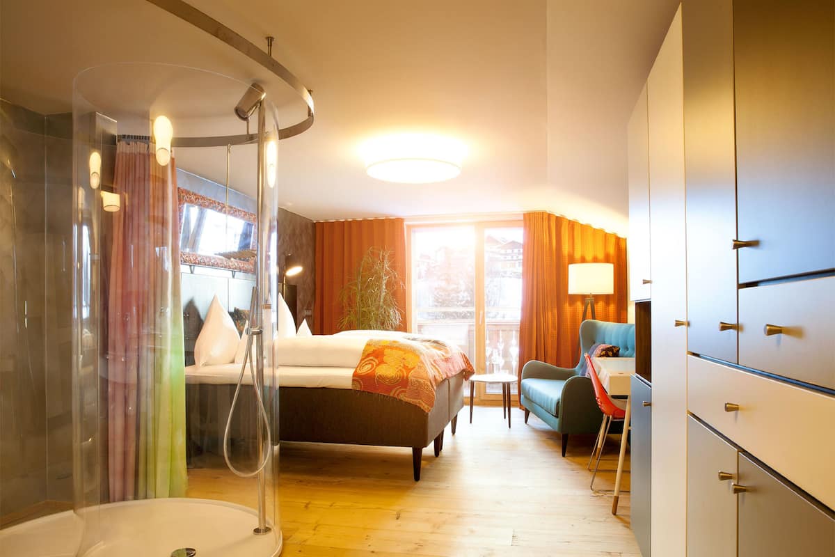 A hotel room with a shower enclosure, a bed with a blanket, a chair, a desk, and a wardrobe.