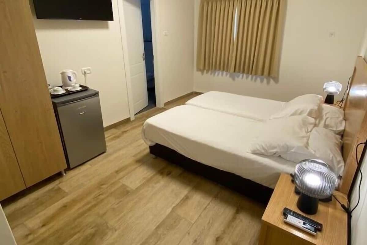 Habitación de hotel con cama, mesita de noche con lámpara, televisor montado en la pared, pequeño escritorio con hervidor y aire acondicionado.