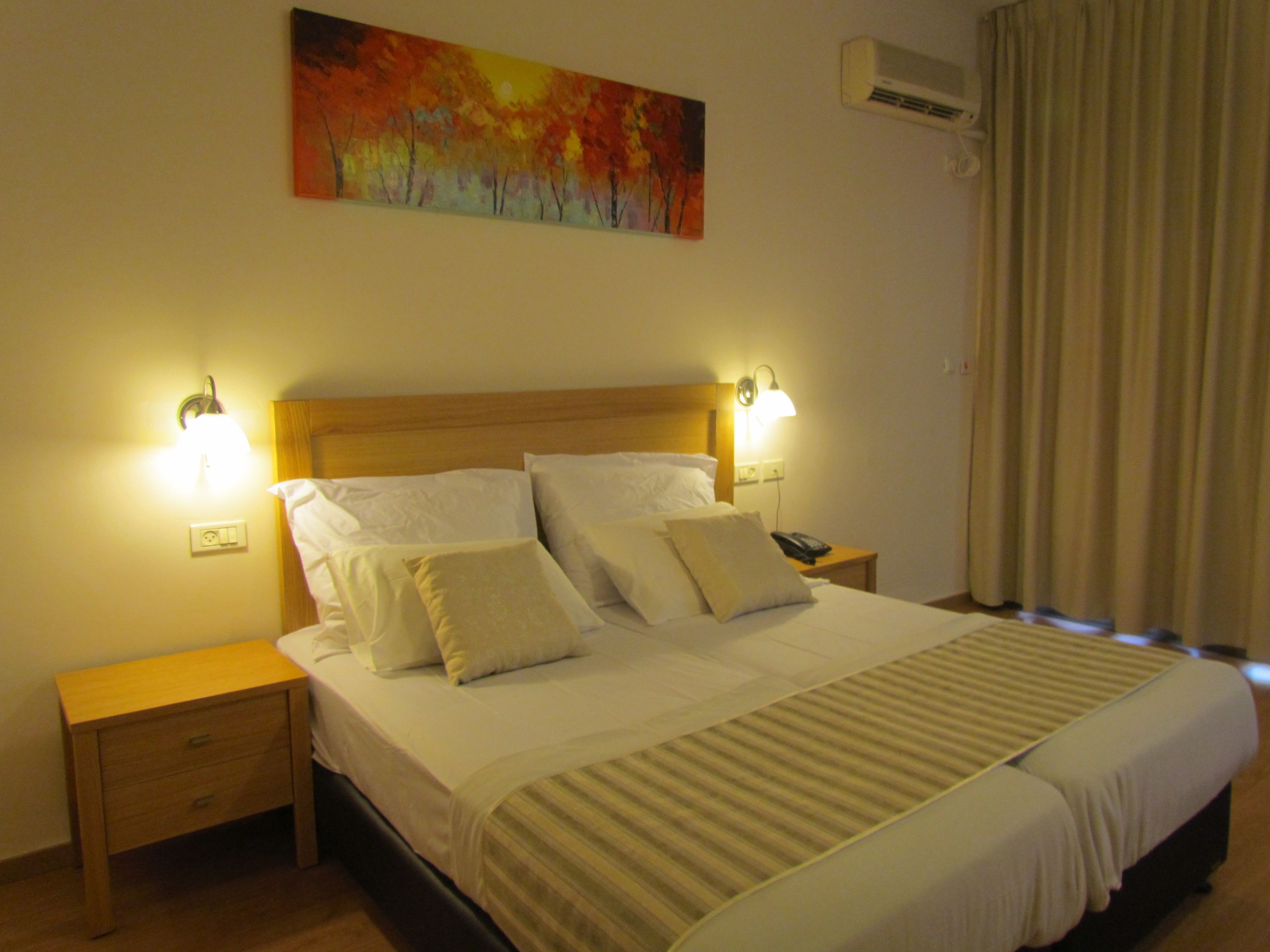 A hotel room with a large bed, two bedside tables, a nightstand, a wall-mounted air conditioner, and a painting above the bed.