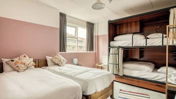 A room with two single beds, a bunk bed, a window with curtains, a bedside table with a lamp, and a striped rug.
