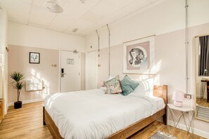 Un dormitorio con cama de madera, ropa de cama blanca, una planta en maceta y una obra de arte enmarcada en la pared.