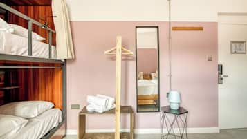A room with bunk beds, a wooden hanger, a mirror, a small table with a lamp, and a door.