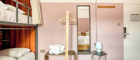 A room with bunk beds, a wooden hanger, a mirror, a small table with a lamp, and a door.