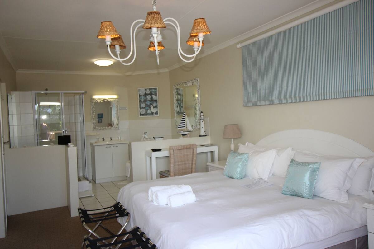 A hotel room with a large bed, a desk, a chair, a mirror, and a bathroom visible through an open door.