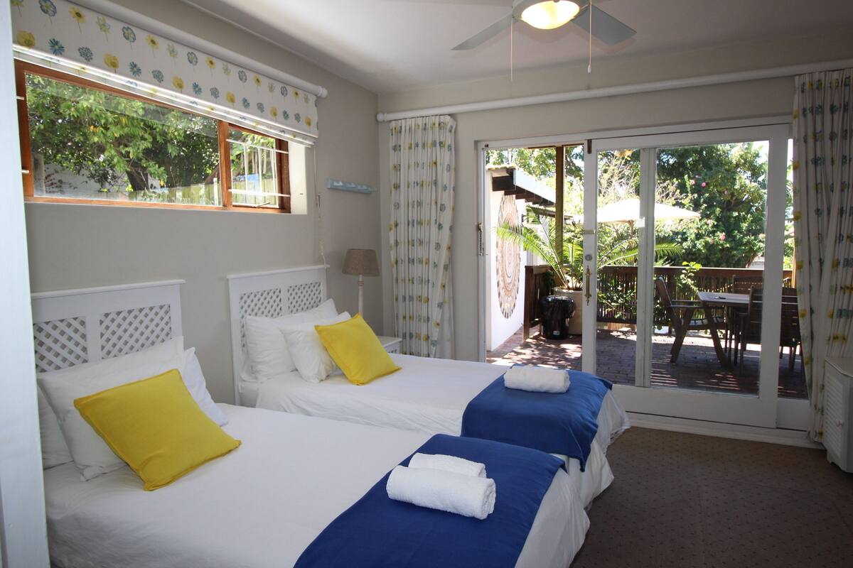A hotel room with two beds, a ceiling fan, and a sliding glass door leading to an outdoor area with a table and chairs.