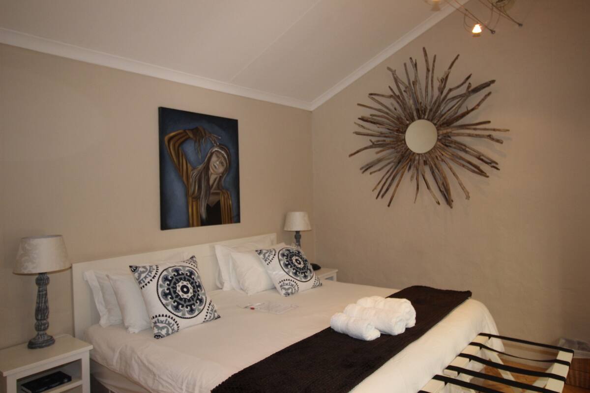 A neatly arranged bedroom with a bed, two bedside tables, a wall-mounted artwork, and a decorative sunburst mirror.