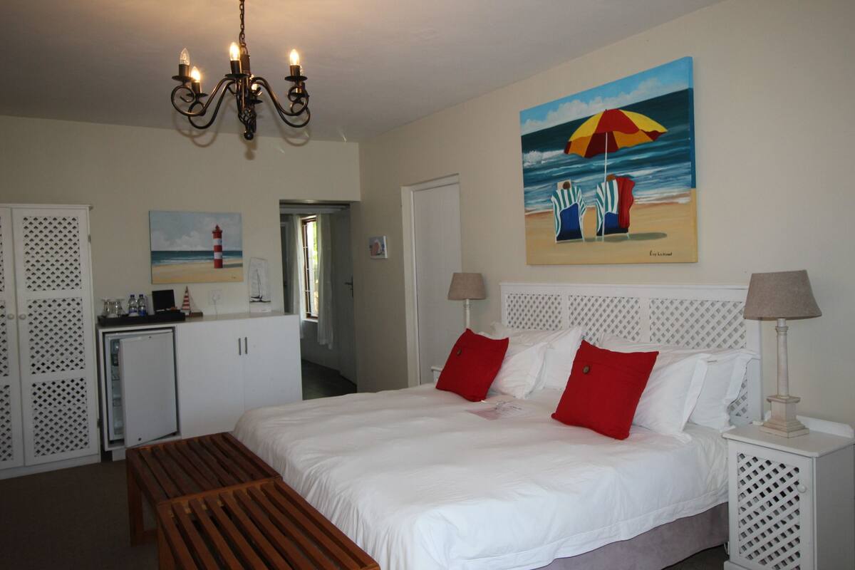 A bedroom with a bed, bedside lamps, a bench, a painting of a beach scene, and a refrigerator.
