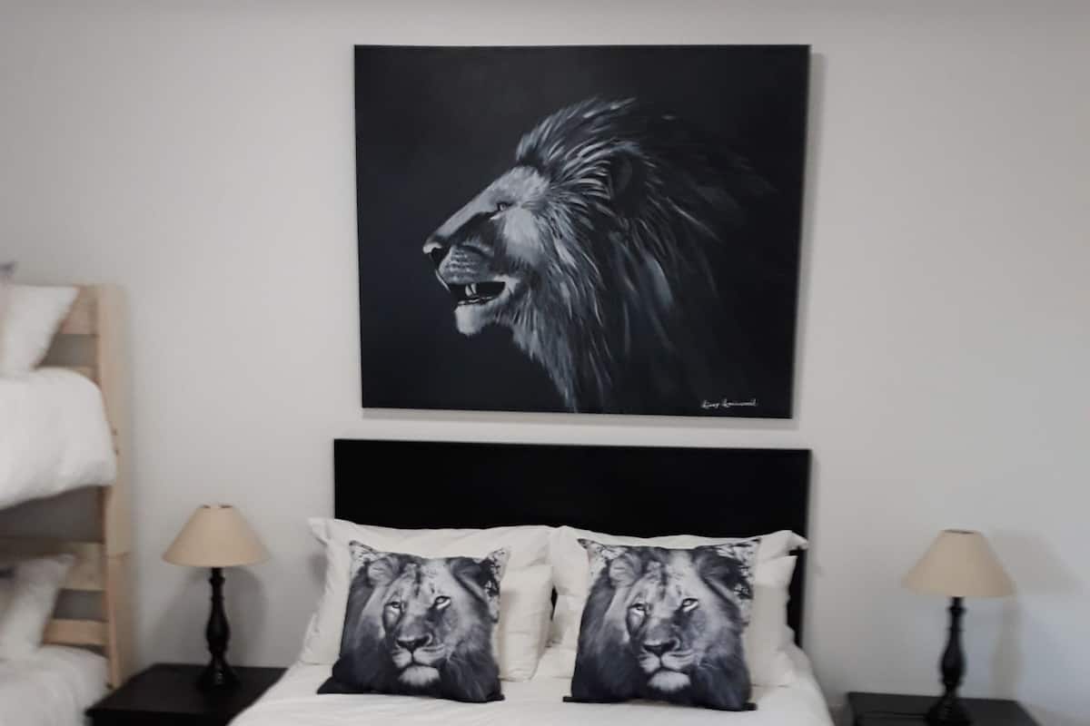 A hotel room with a large bed, two pillows with lion prints, a black headboard, two bedside tables with lamps, and a wooden ladder against the wall.