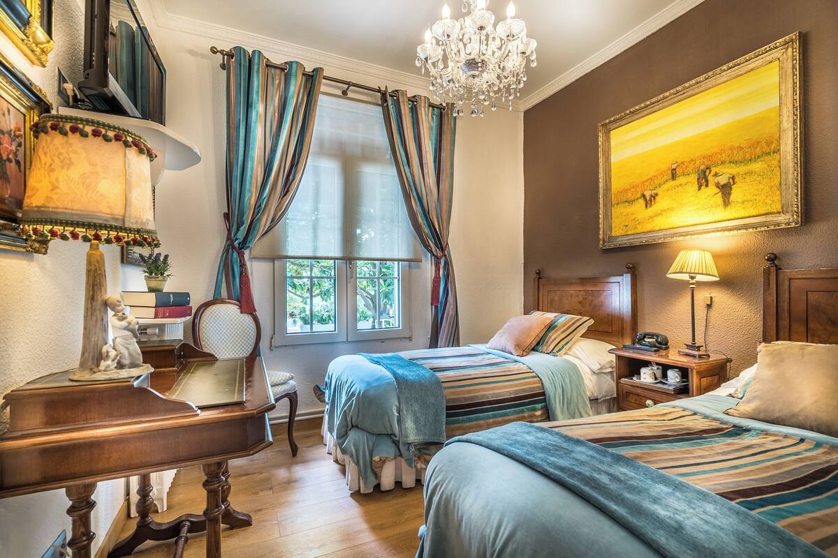 A bedroom with two beds, a chandelier, a painting, and a desk with a lamp.