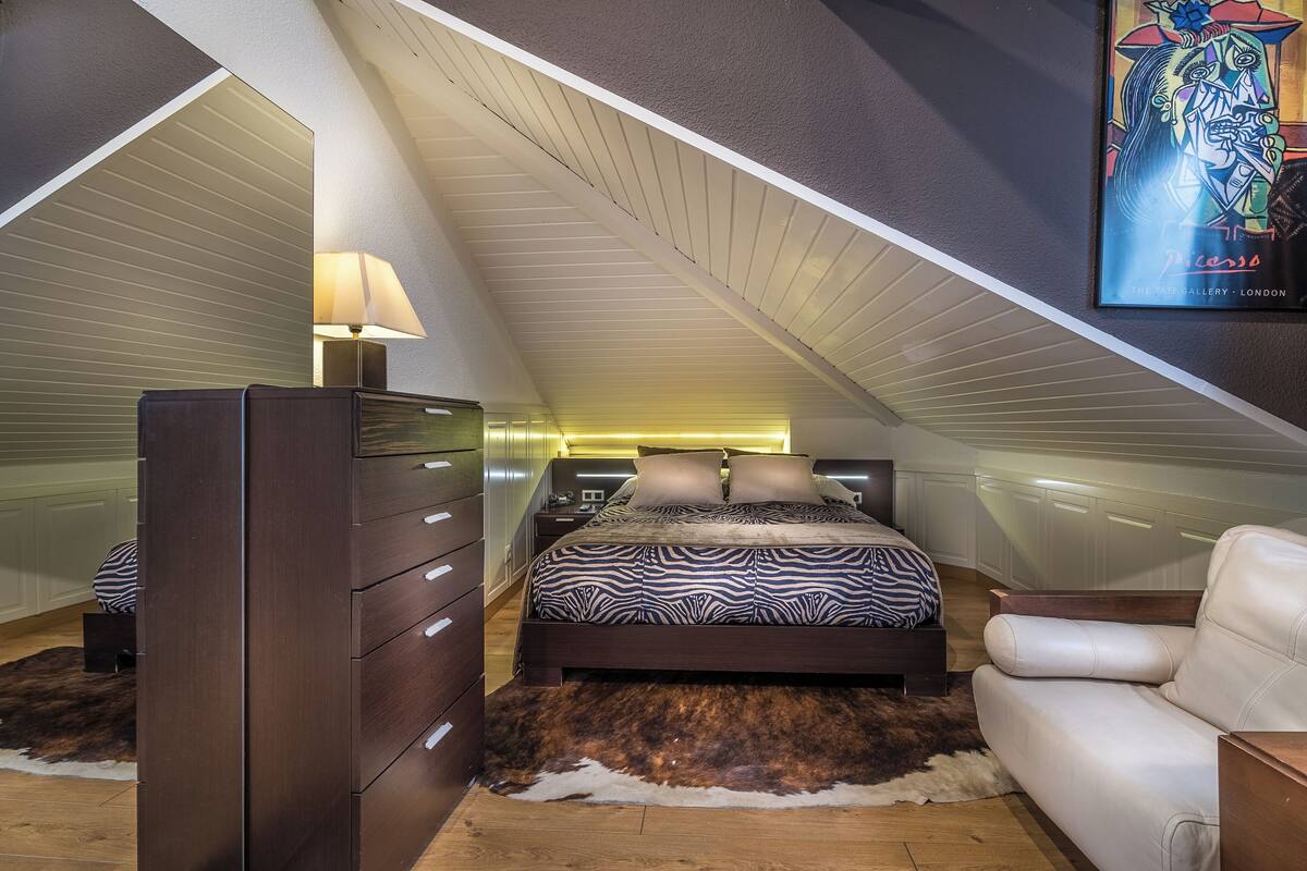 A bedroom with a sloped ceiling, a bed with zebra-patterned bedding, a dresser, a white leather sofa, and a framed artwork on the wall.