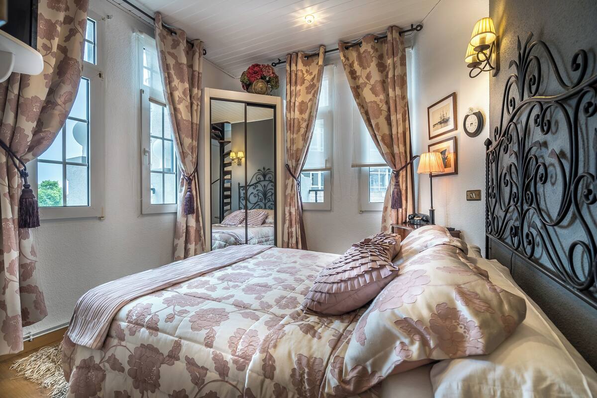 A bedroom with a large bed, floral curtains, a mirror, and wall-mounted lamps.