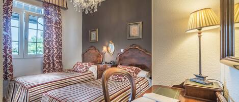 A bedroom with two beds, a chandelier, a desk with a lamp, and framed pictures on the wall.