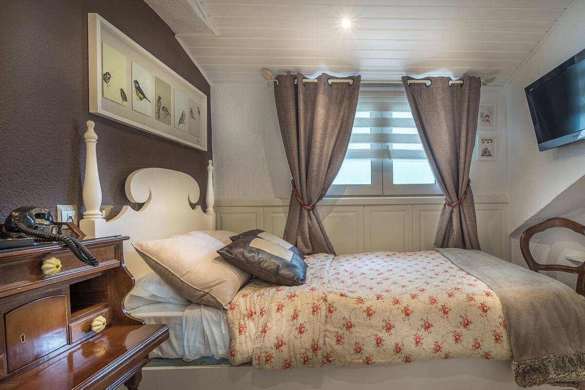 A bedroom with a bed, a bedside table with a telephone, a television mounted on the wall, and a window with curtains.