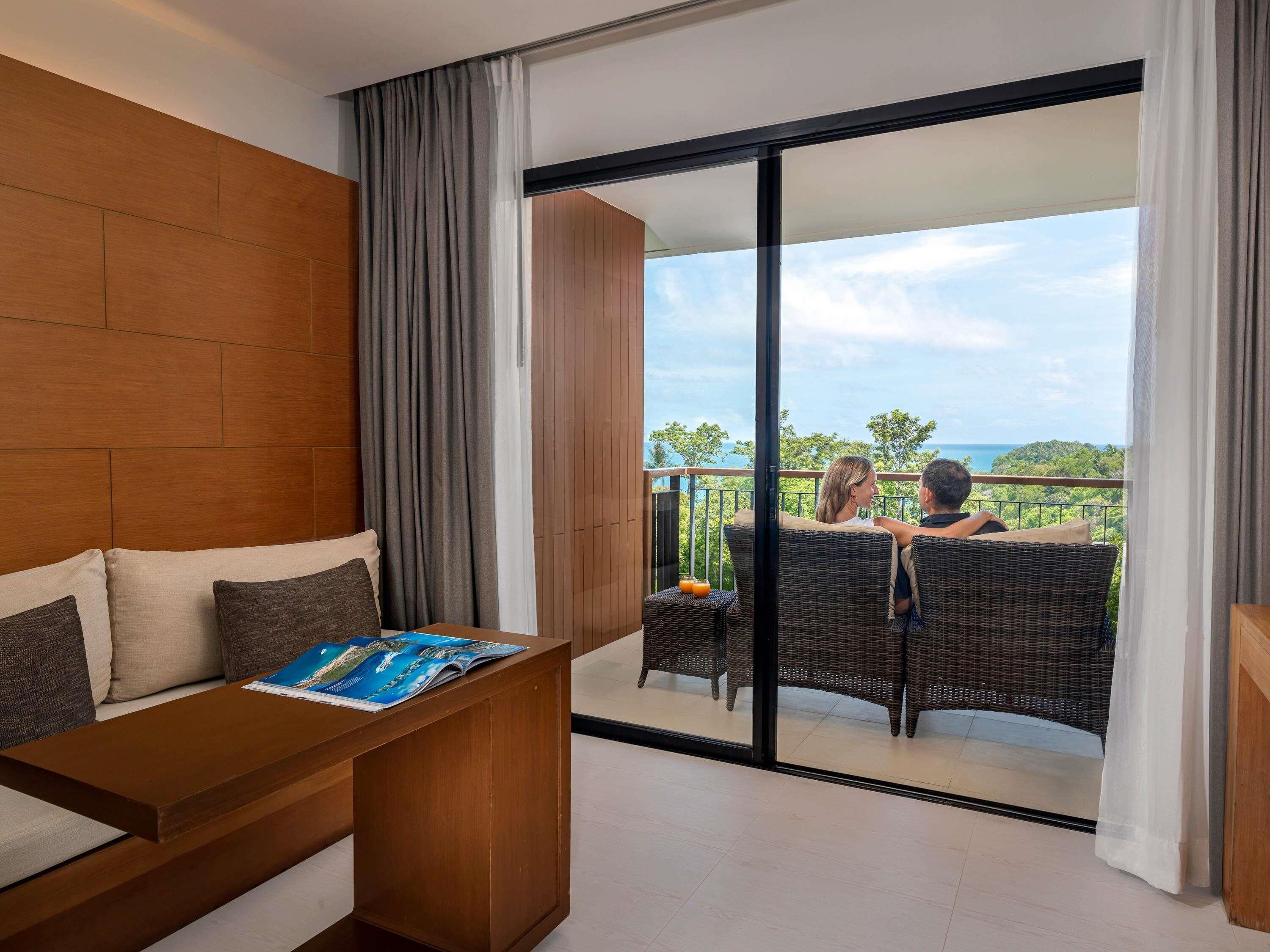 A modern hotel room with a wooden headboard, a wooden table with a magazine, and a balcony with wicker furniture overlooking a scenic view.