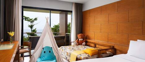 Habitación de hotel con cama, cabecera de madera, una mesita con un florero, un sillón de peluche azul, un osito de peluche y vista al océano.