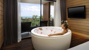Ein Zimmer mit einer großen Badewanne voller Wasser und Rosenblüten, einem an der Wand befestigten Fernseher und einem Balkon mit Meerblick.