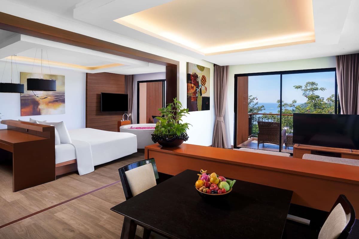 Een moderne hotelkamer met een bed, een bureau, een eettafel met fruit, een televisie en een balkon met uitzicht.