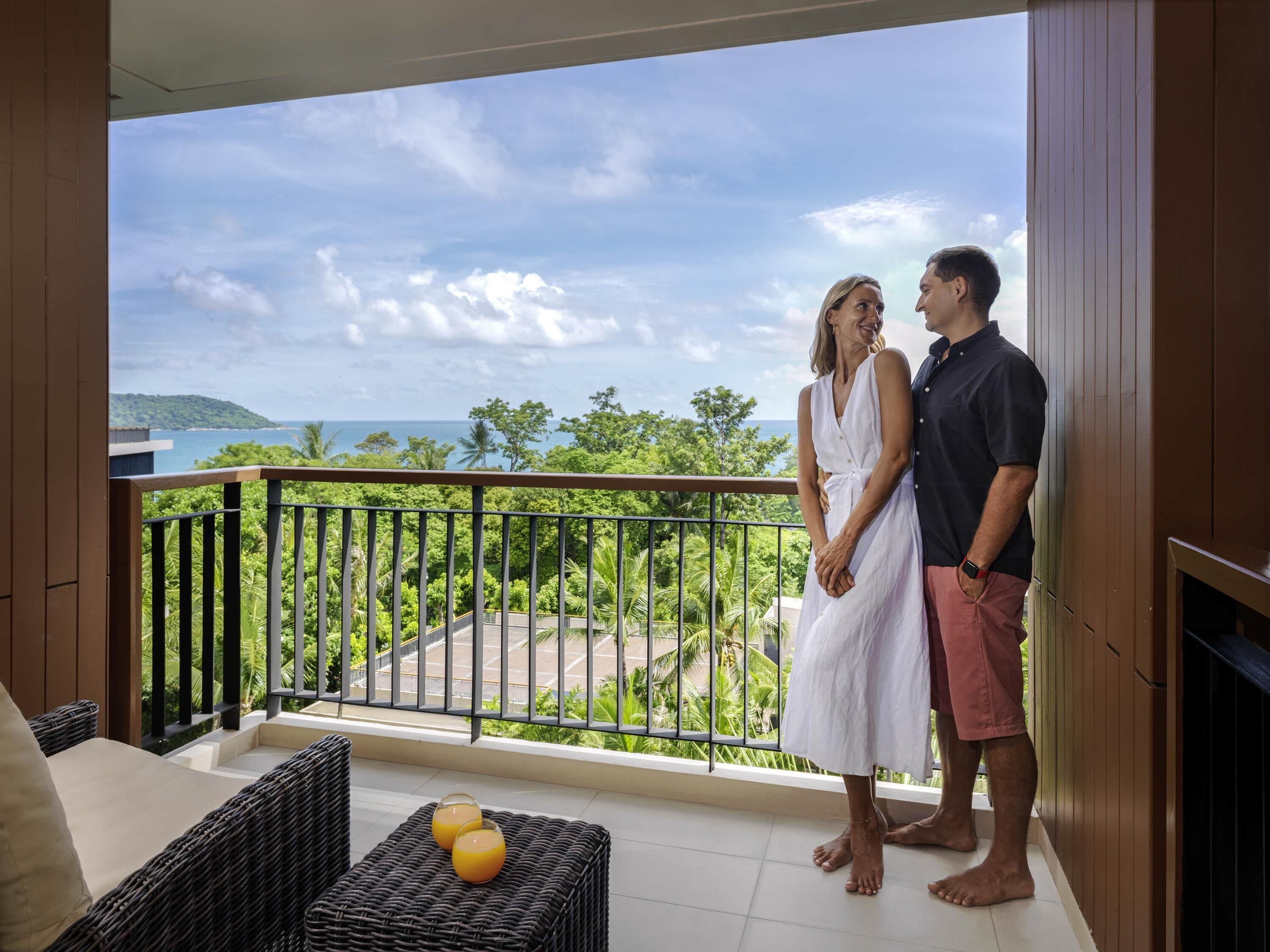 A balcony with a view of the ocean and greenery, wicker furniture, and two people standing.