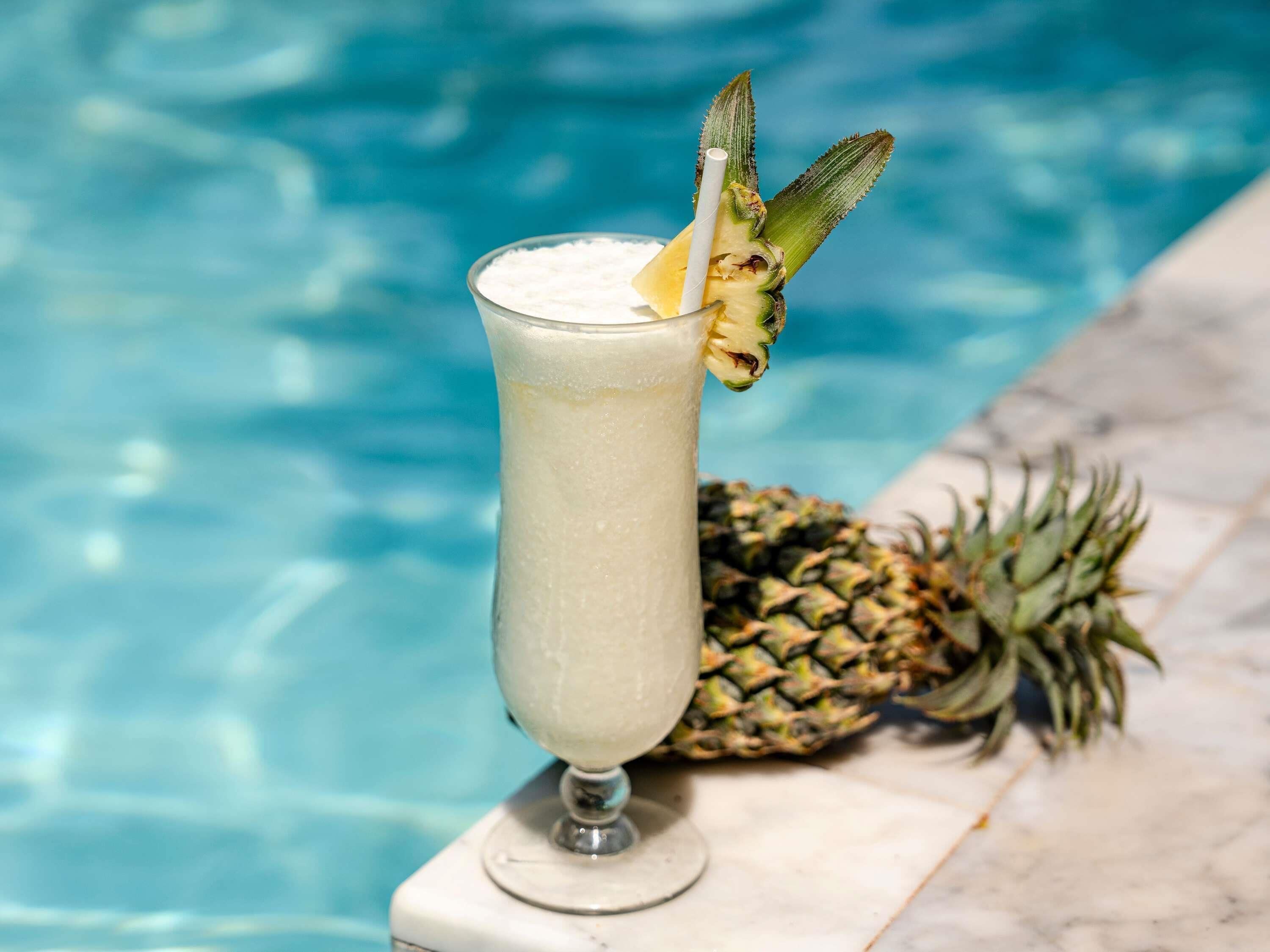 A glass of a light-colored beverage with a pineapple garnish, placed on a marble surface near a pool.