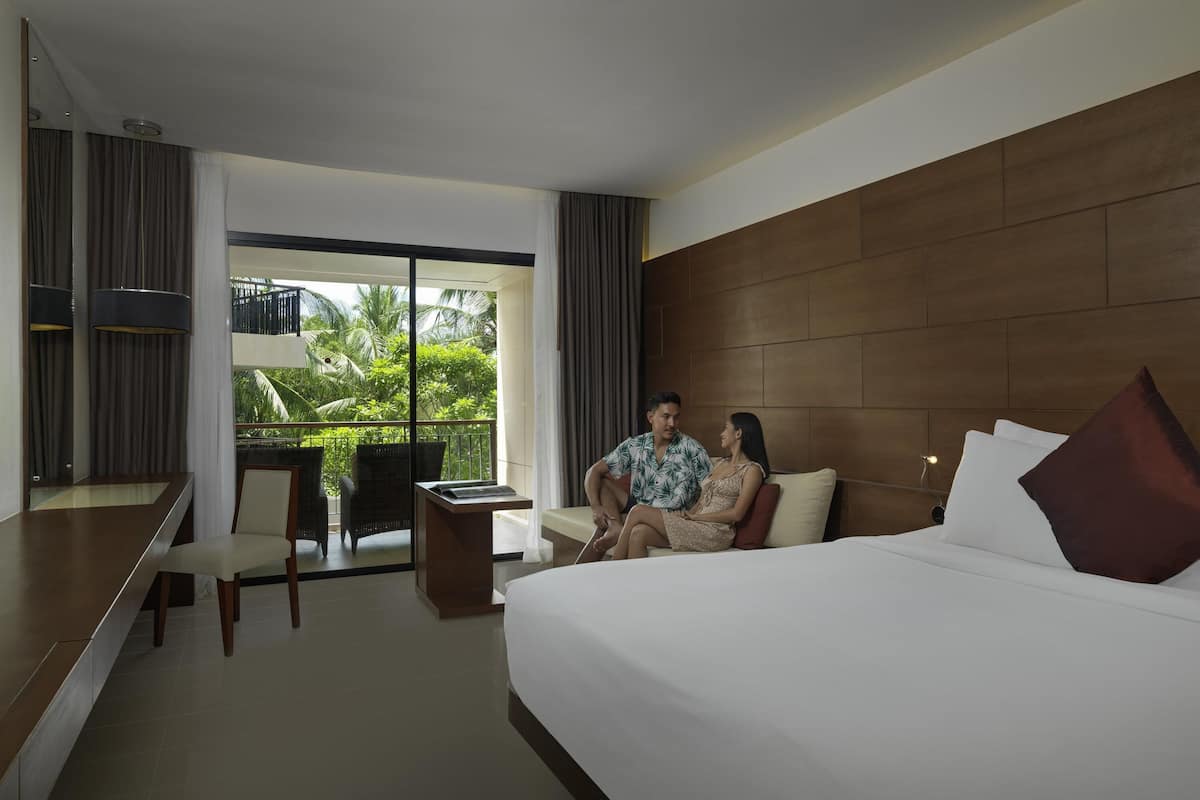 Een hotelkamer met een groot bed, een bureau, een stoel en een balkon met uitzicht op groen.