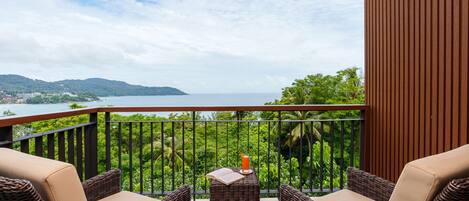 A balcony with wicker furniture and a table, overlooking a lush landscape and the sea.