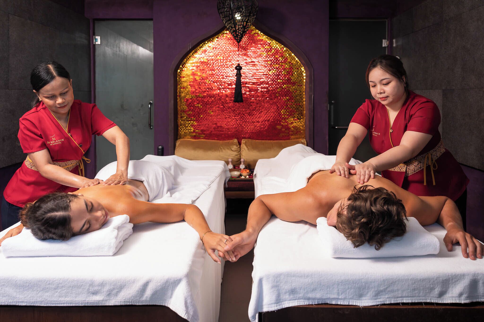 Two massage tables with clients receiving back massages from two therapists in red uniforms.