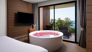 A modern hotel room with a large bathtub filled with rose petals, a flat-screen TV mounted on the wall, and a balcony with outdoor furniture.