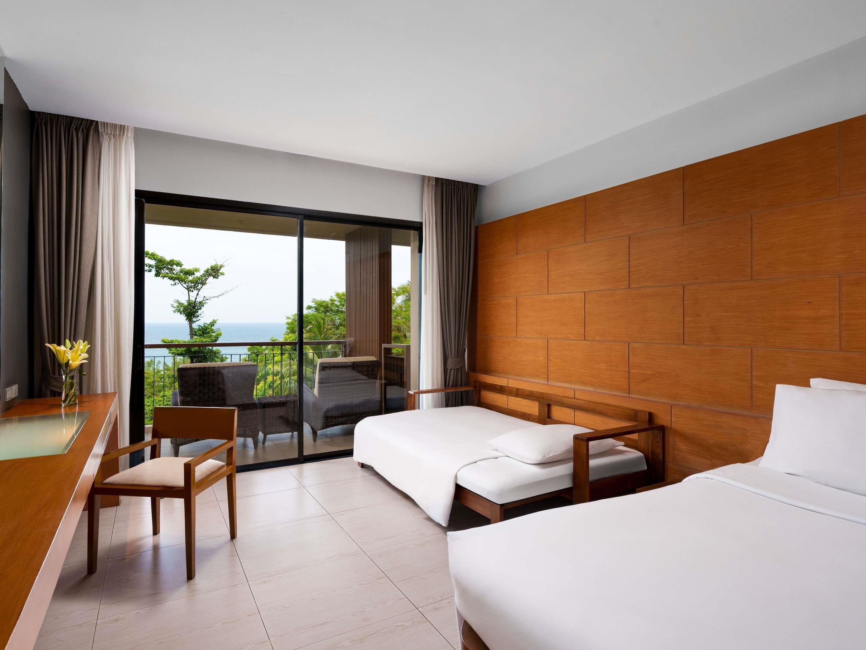 A hotel room with a balcony, a desk, two beds, and a view of the sea.