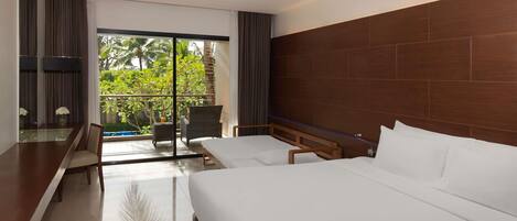 A hotel room with a large bed, a desk, a chair, and a balcony with a view of greenery.