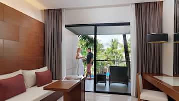 A modern hotel room with a wooden headboard, a sofa with red cushions, a glass-top table, a wicker chair, and a view of a pool and palm trees through a sliding glass door.