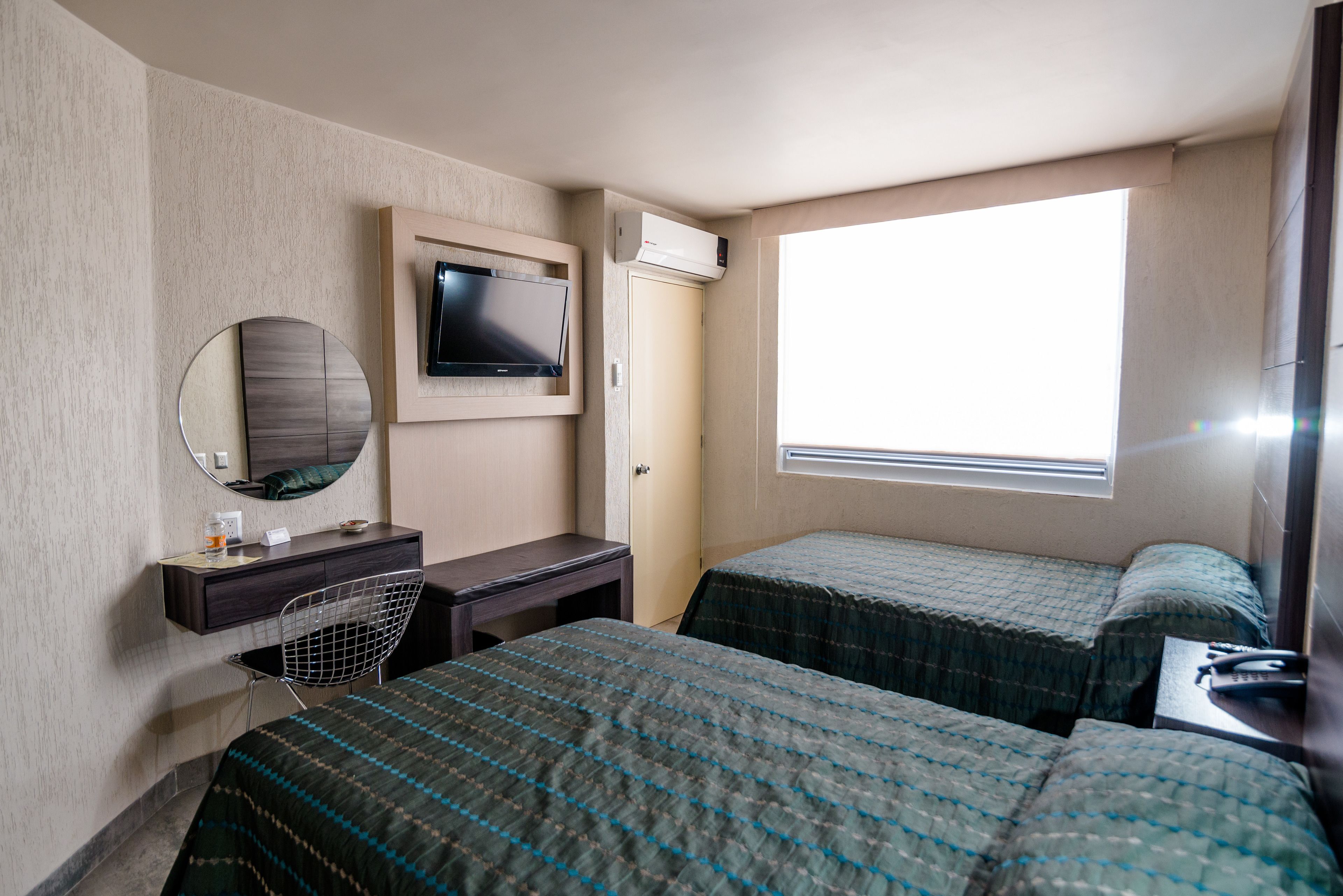 A hotel room with two beds, a desk with a chair, a television, and a window providing natural light.