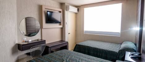 A hotel room with two beds, a desk with a chair, a television, and a window providing natural light.