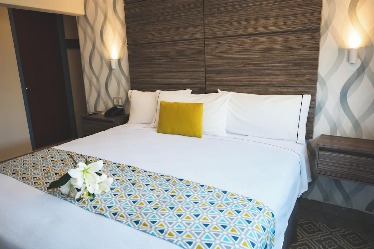 Habitación de hotel con una cama bien hecha, una mesita de noche con lámpara y una colcha estampada.
