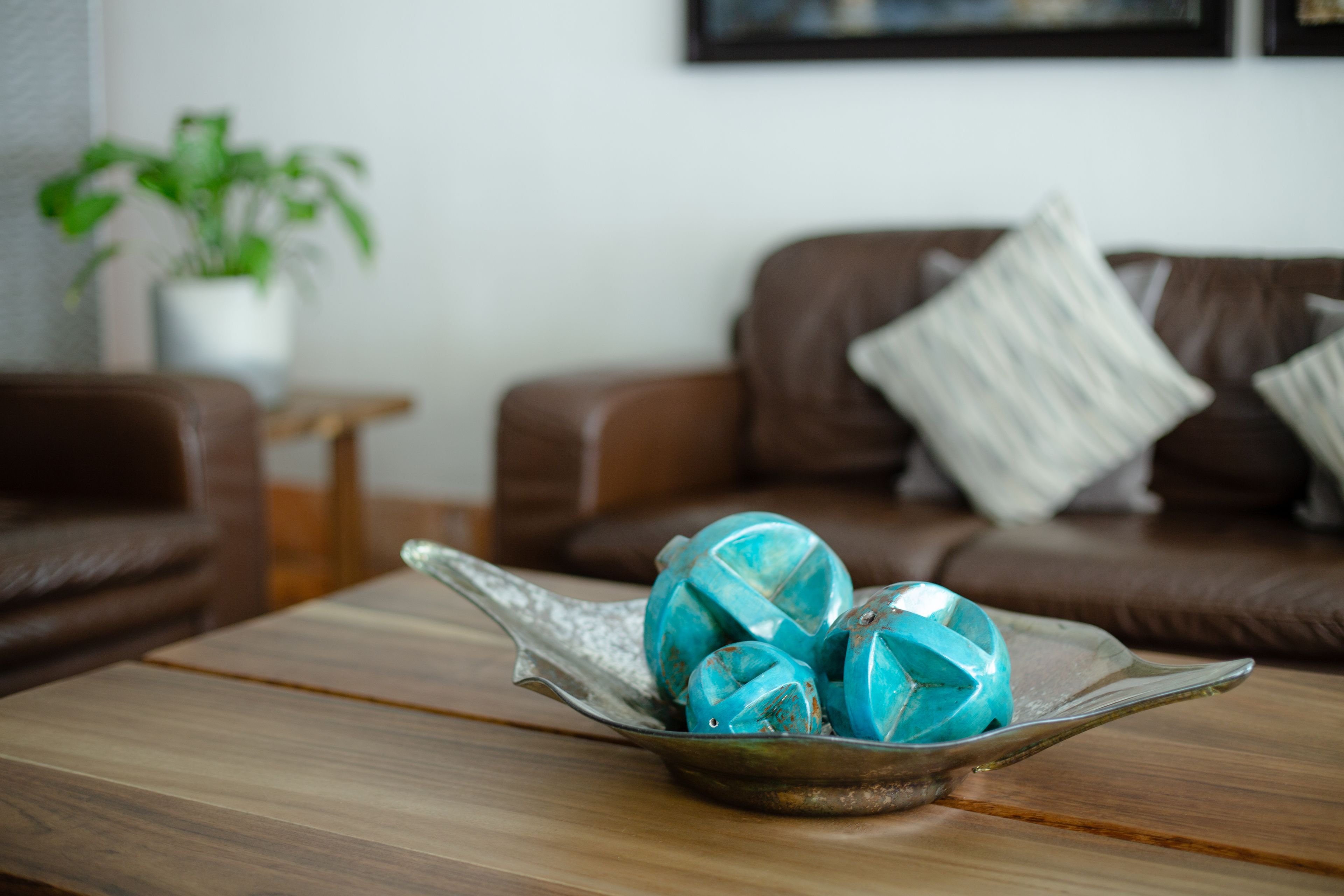 A living room with a brown leather sofa, a small wooden side table, and a glass bowl containing turquoise ornaments on a wooden coffee table.