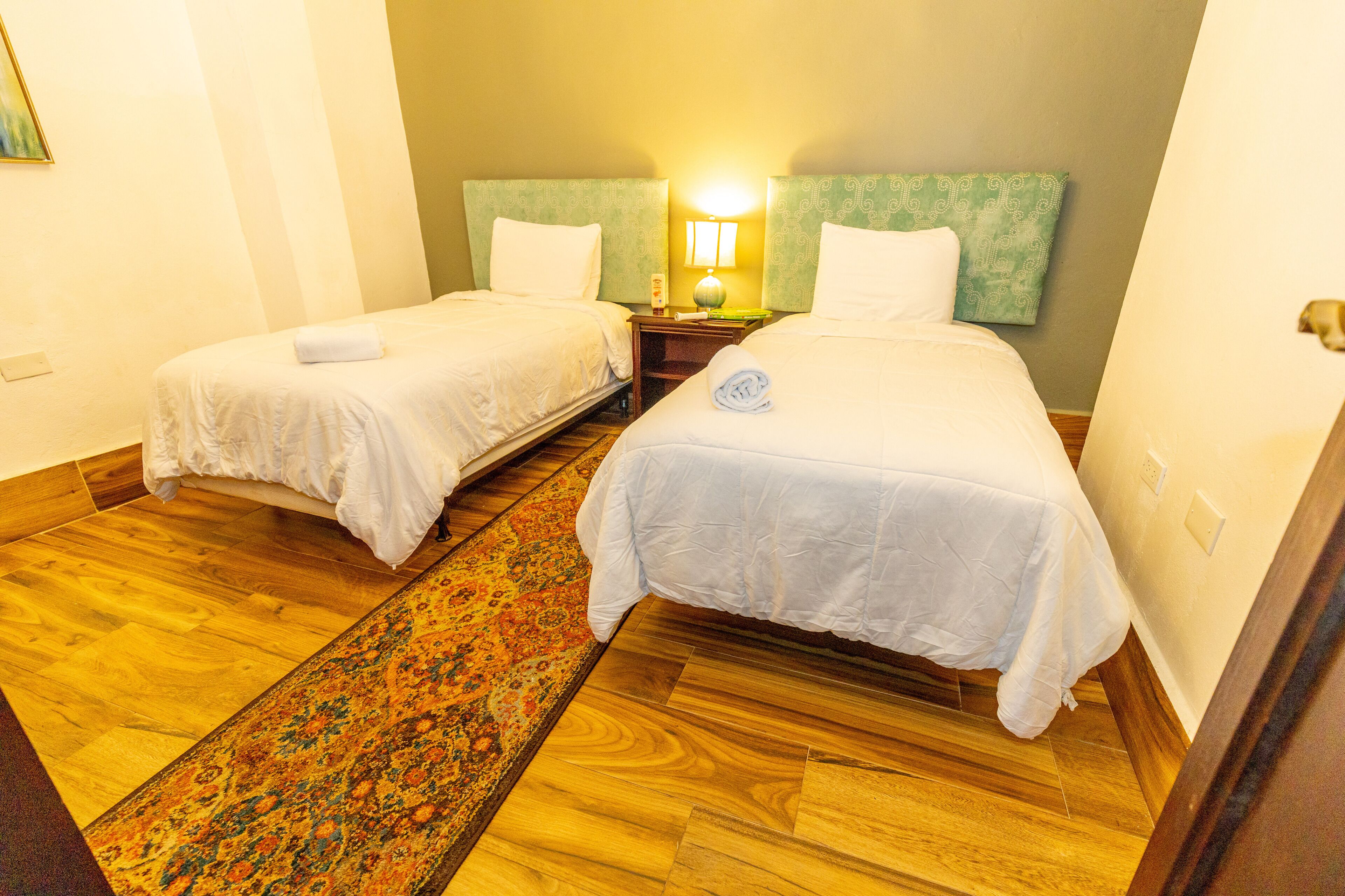 A hotel room with two single beds, a nightstand with a lamp, a patterned rug, and wooden flooring.
