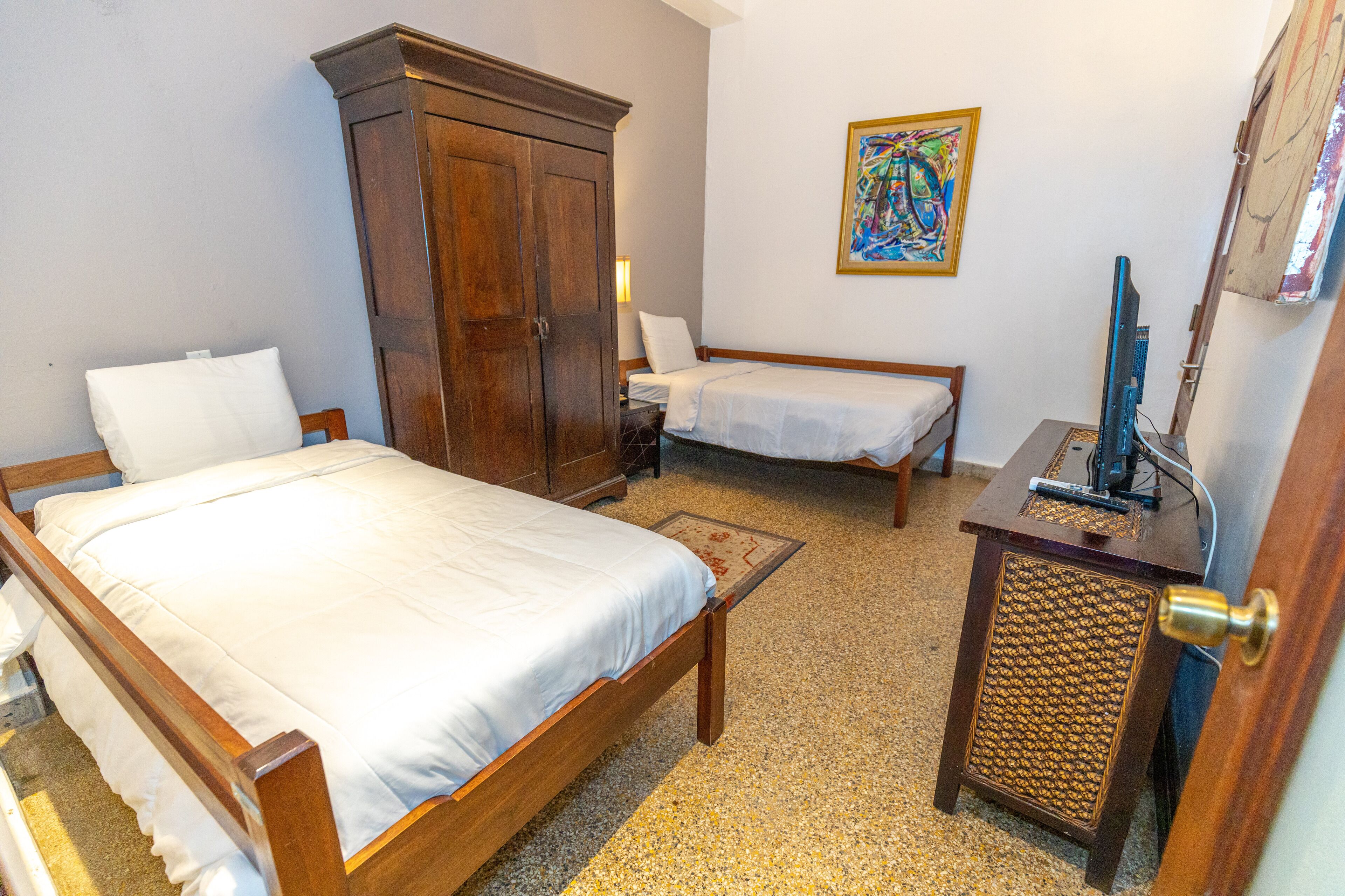 A bedroom with two single beds, a wooden wardrobe, a small table with a speaker, and a painting on the wall.