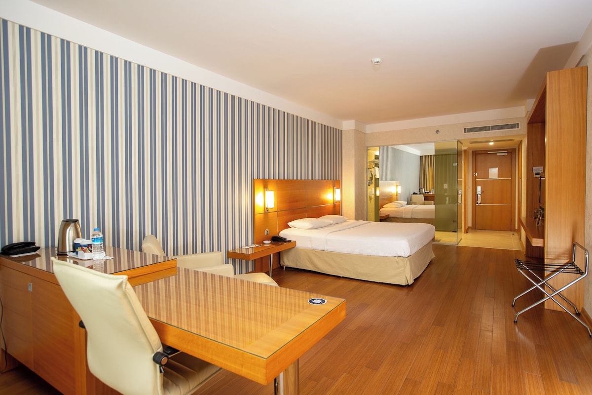 A hotel room with a bed, a desk, and a chair. There is a mirror on the wall, and a door leading to a bathroom.