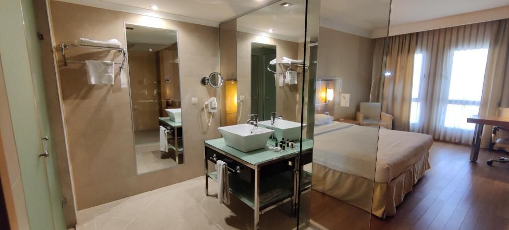 A hotel room with a large bed, two bedside tables, a double sink, a mirror, and a shower area.