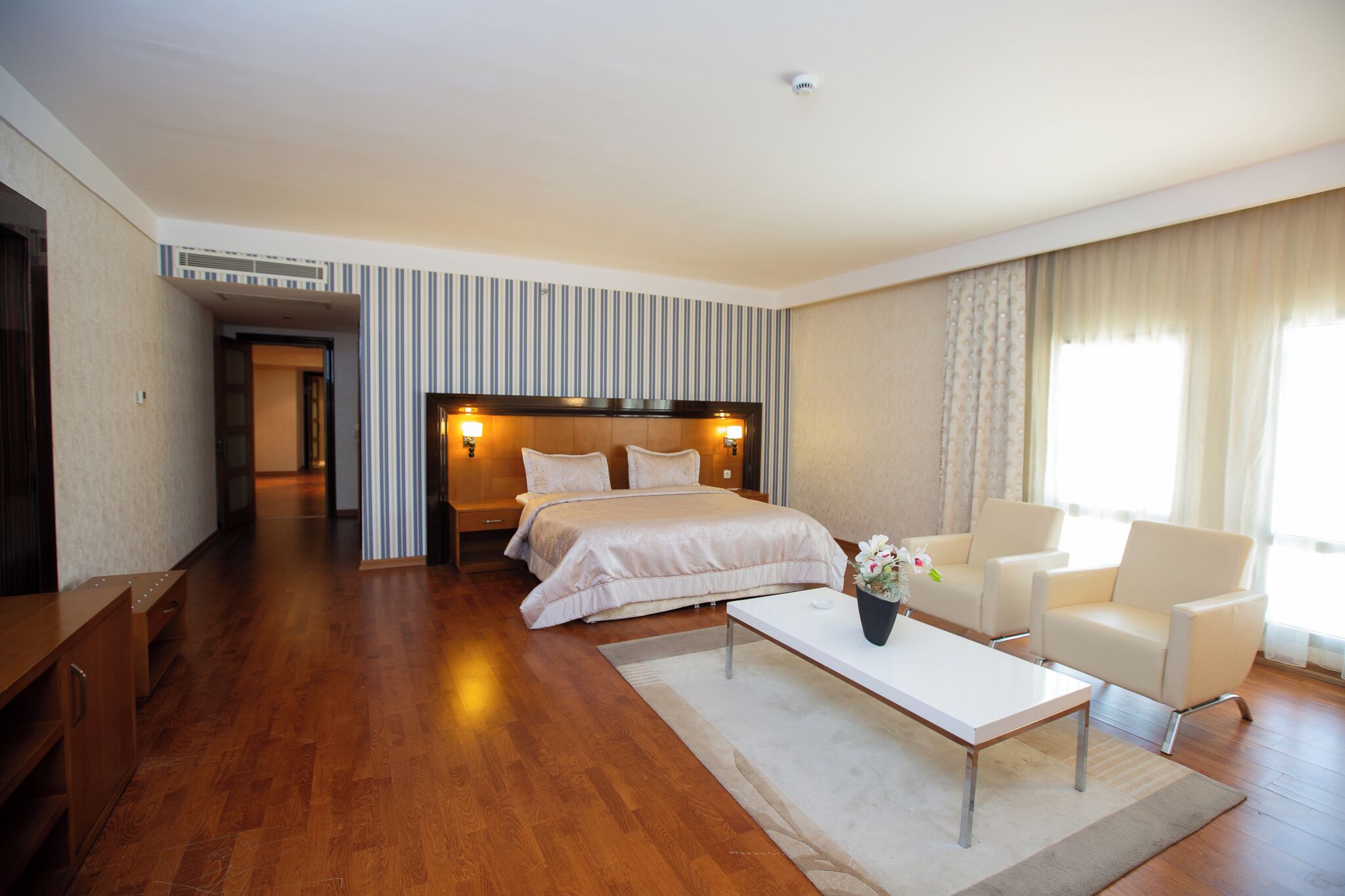 Een hotelkamer met een groot bed, nachtkastjes, een zithoek met stoelen, een klein tafeltje en een raam met gordijnen.