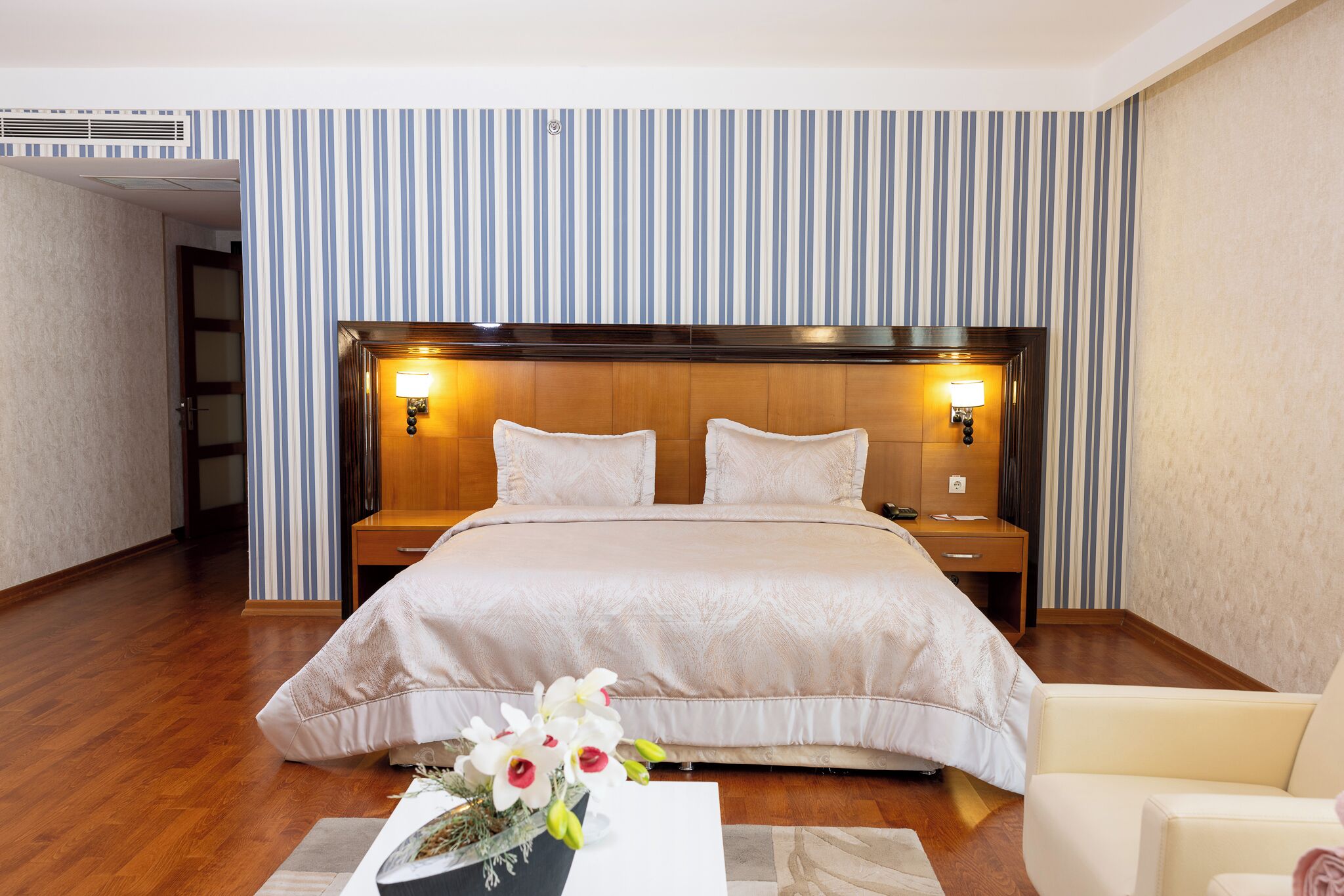 A hotel room with a large bed, two bedside tables, a nightstand, a chair, and a vase of flowers on a coffee table.
